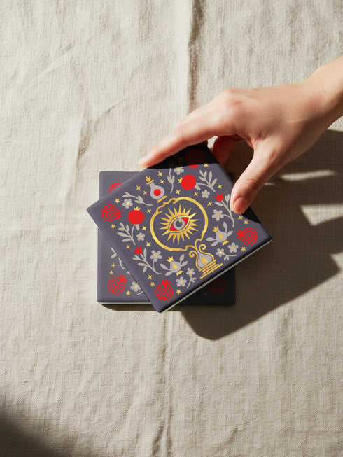 A square ceramic drink coaster with a smooth, matte charcoal grey finish. The surface features a symmetrical, intricate design in metallic gold, deep garnet red, and light grey. In the center is a golden All-Seeing Eye with a red iris, radiating light. The eye is framed by a golden circle and flanked by botanical elements, including red pomegranates and grey flowers with gold details. The design has a folk art, gothic botanical style.